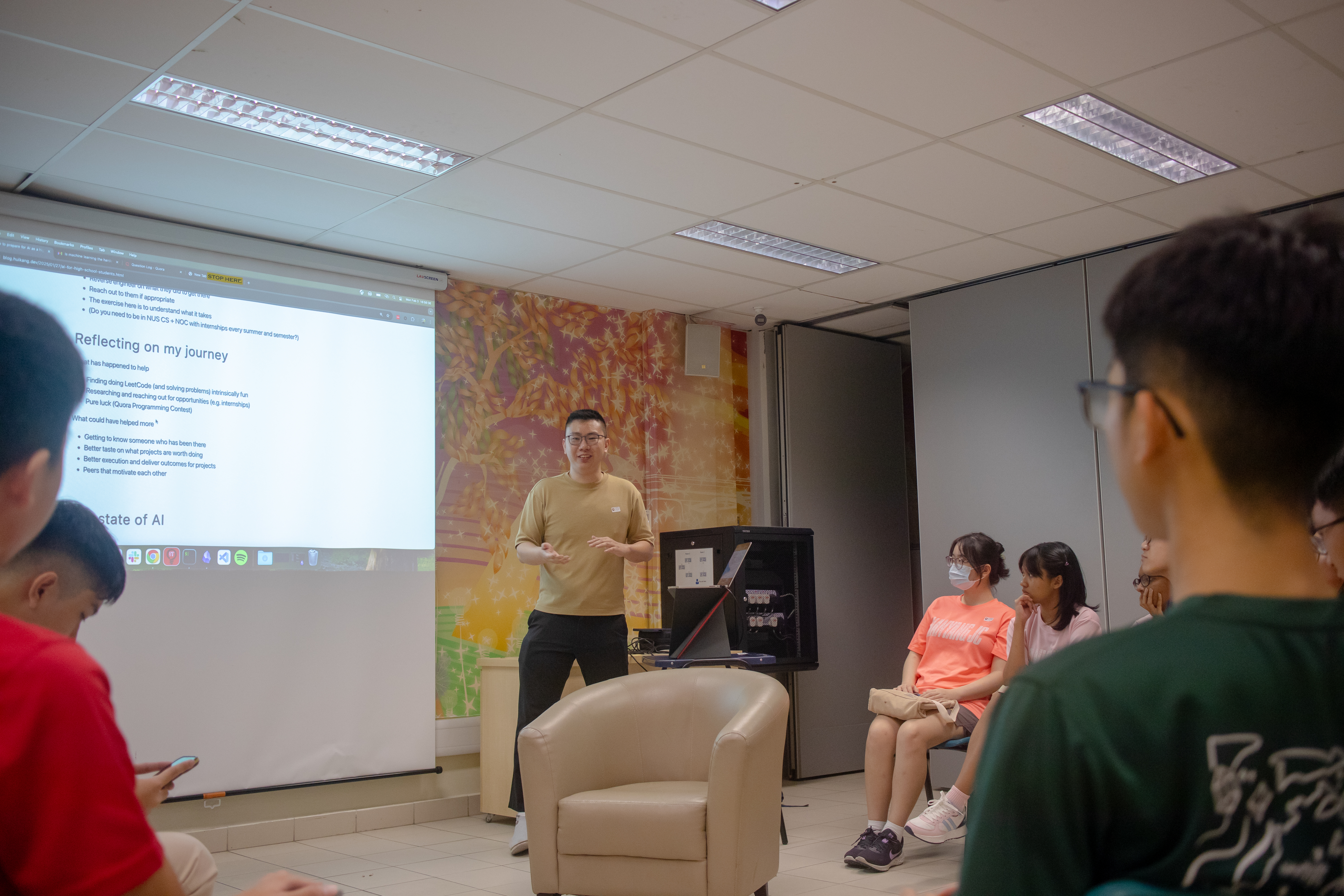 A speaker, Huikang, standing at the front of a classroom next to a presentation slide titled 'Reflecting on my journey.' Students are seated attentively, with a cozy setup featuring chairs and bright walls.